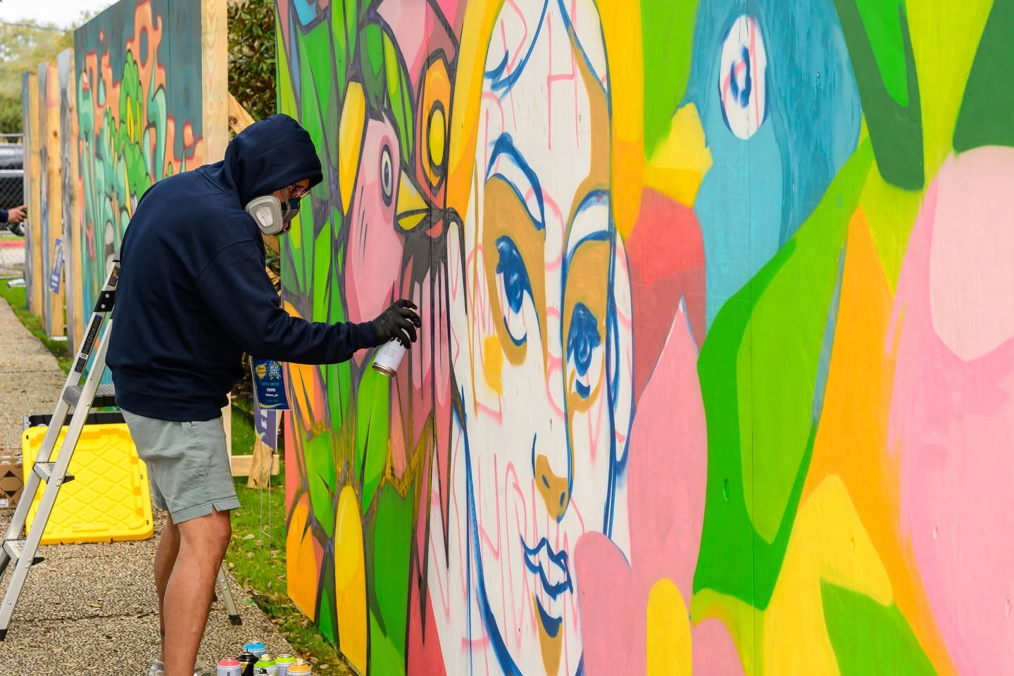 Artist sprays colorful mural on a wooden fence, wearing mask and gloves.